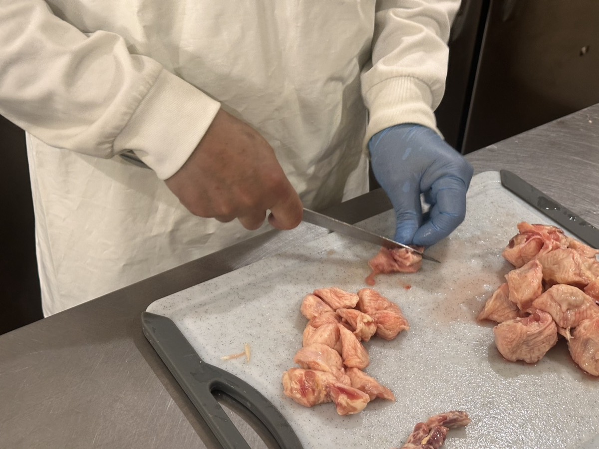 白衣と手袋の職人が、まな板の上で鶏肉を包丁で切り分けている手元。